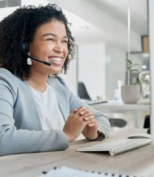 Smiling virtual receptionist wearing a headset, sitting at a desk with a computer, providing customer support in an office environment.