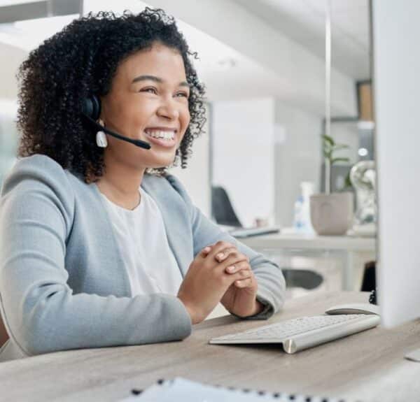 Smiling virtual receptionist wearing a headset, sitting at a desk with a computer, providing customer support in an office environment.