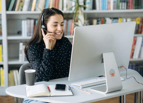 Professional Virtual Receptionist at Work A smiling woman wearing a headset works on a desktop computer in a professional setting with bookshelves in the background.