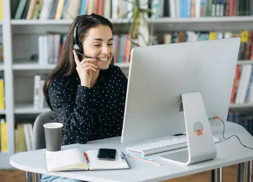 A smiling woman wearing a headset works on a desktop computer in a professional setting with bookshelves in the background.