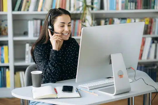 A smiling woman wearing a headset works on a desktop computer in a professional setting with bookshelves in the background.