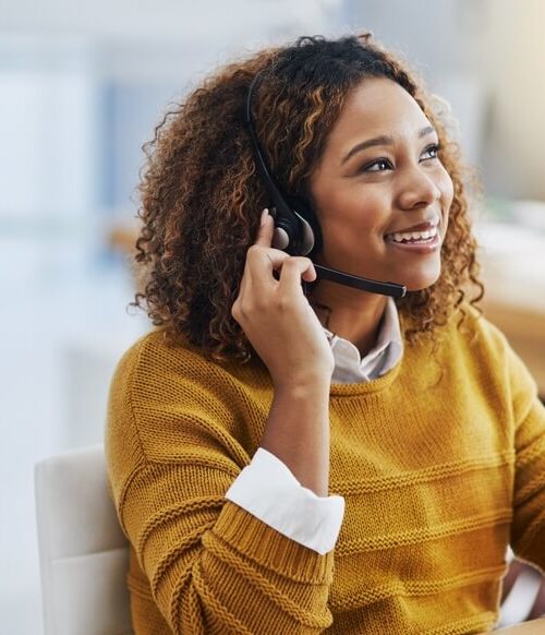 A smiling woman in a mustard sweater provides live answering services while wearing a headset and working on a computer.