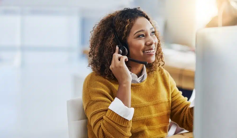 A smiling woman in a mustard sweater provides live answering services while wearing a headset and working on a computer.