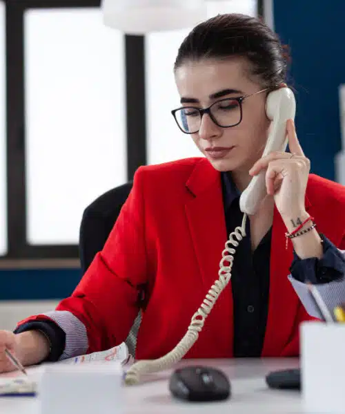 Professional receptionist speaking on a landline phone while writing notes at her desk in a modern office setting