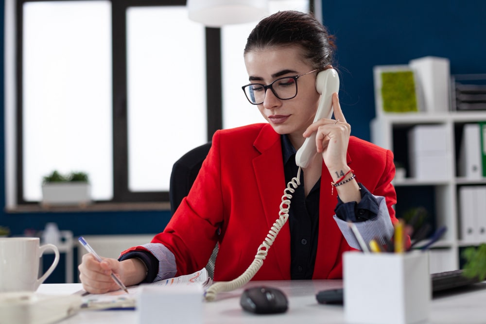 in-house receptionist Professional receptionist speaking on a landline phone while writing notes at her desk in a modern office setting