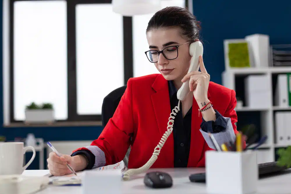 Professional receptionist speaking on a landline phone while writing notes at her desk in a modern office setting
