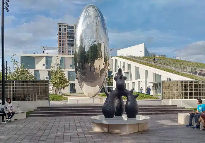 Large reflective sculpture in a public park with visitors walking around.