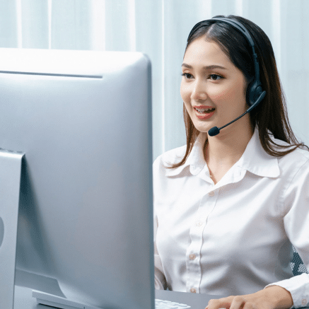 A virtual receptionist wearing a black headset with a microphone sits at a desk in front of a large desktop computer monitor, typing on a keyboard.