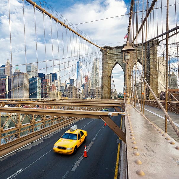 Brooklyn Bridge with yellow taxi driving under it, cityscape in the background.